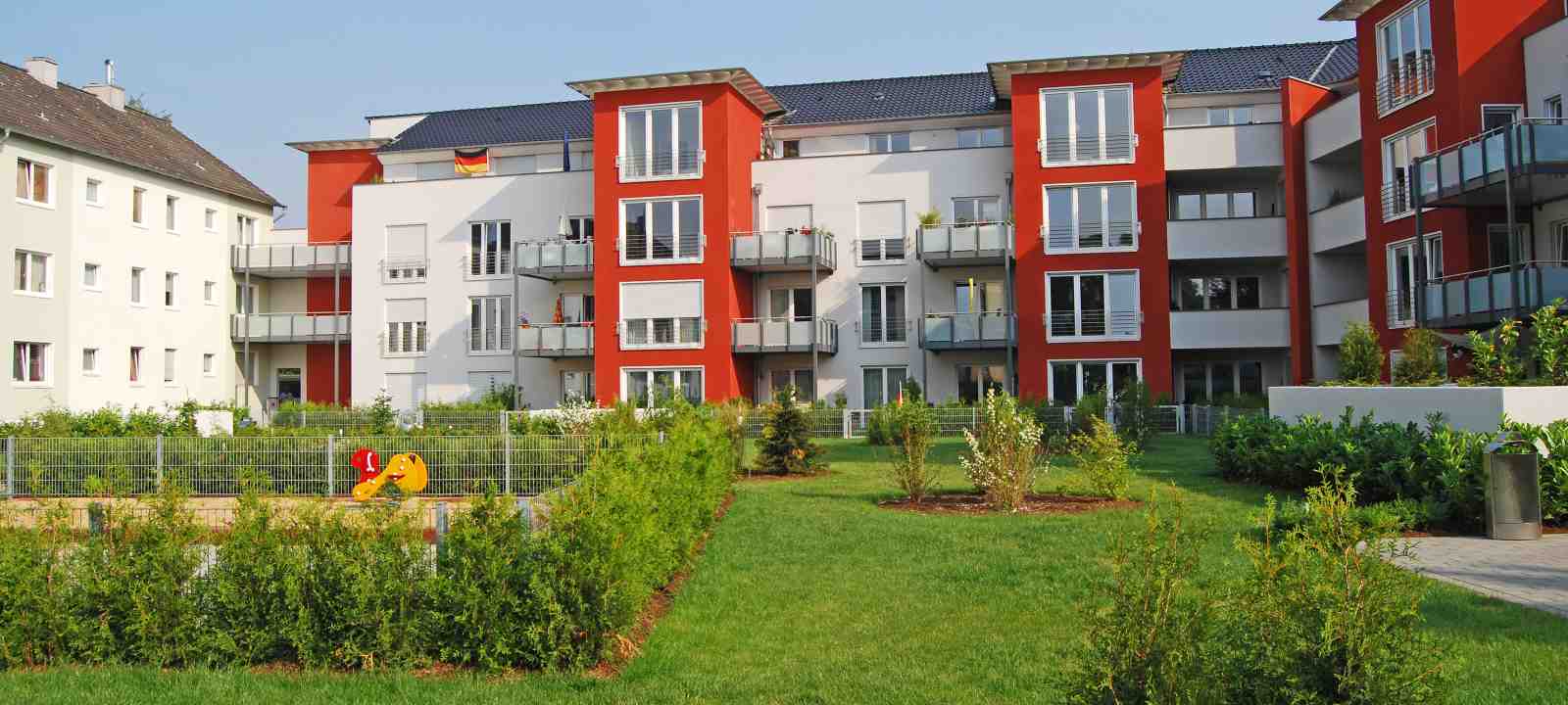 Wohnanlage mit rot-weißen Häusern, gruppiert um eine Rasenfläche mit Sträuchern und Spielplatz